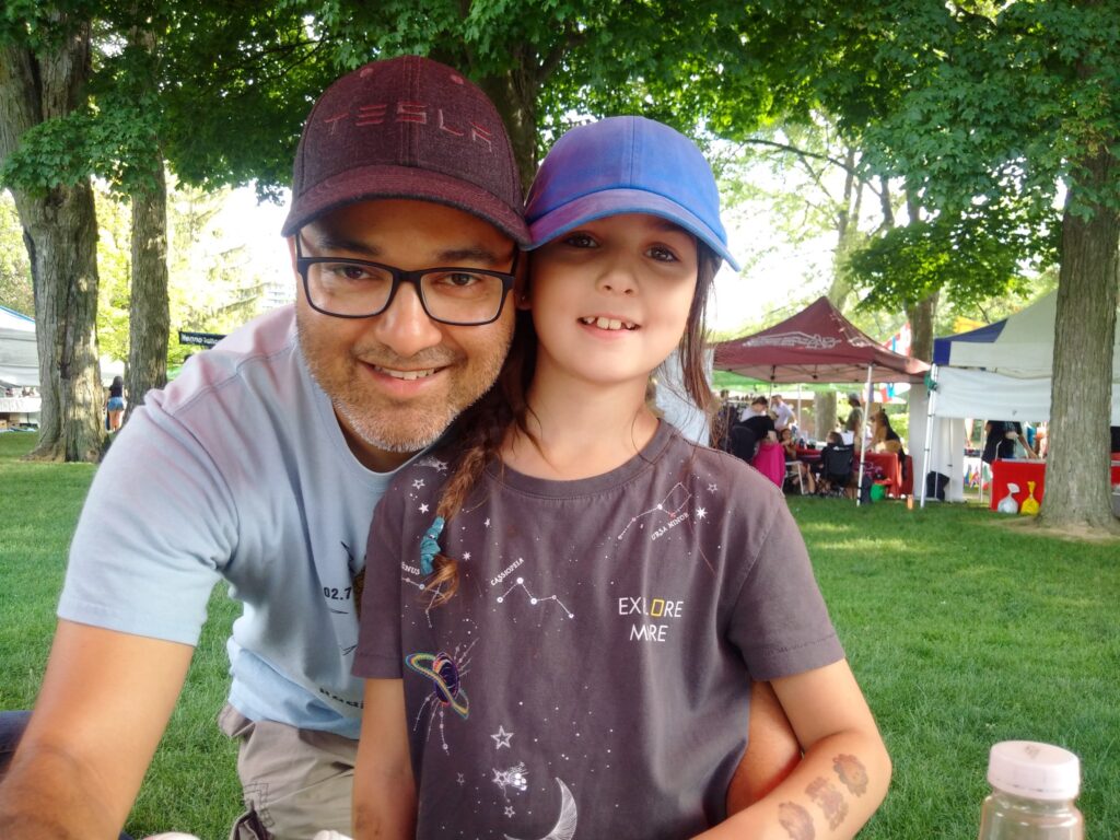 Nat Persaud wearing a CKMS-FM T-shirt and black baseball cap with "Tesla" embroidered on it, and DJ Daisy wearing a blue baseball cap and dark T-shirt, both looking at the camera