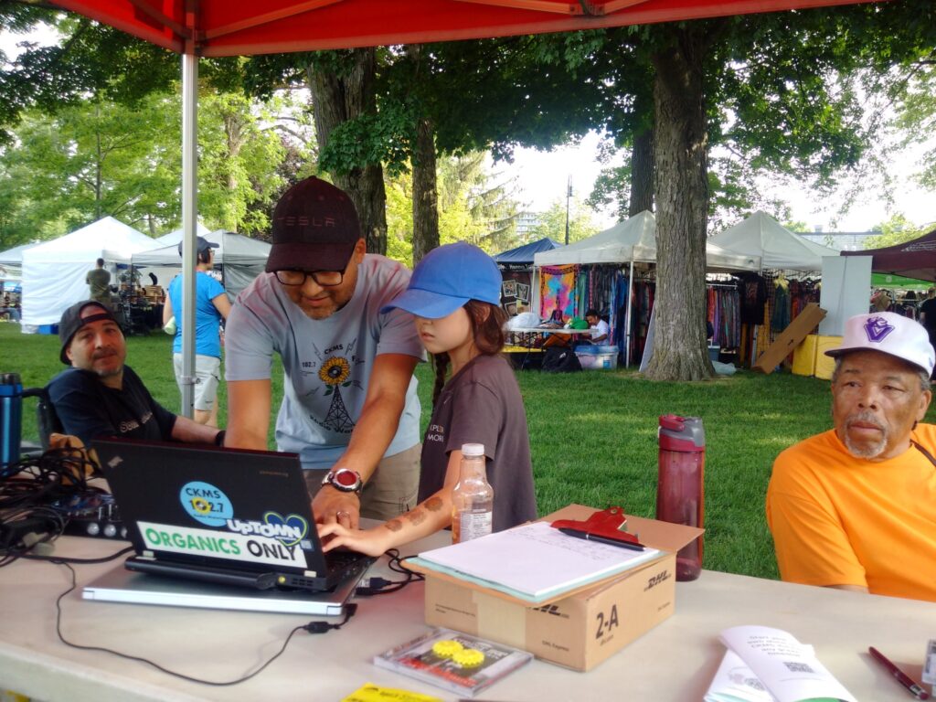 Nat Persaud wearing a black baseball cap and a CKMS-FM T-shirt and DJ Daisy wearing a blue baseball cap and a dark T-shirt work on a laptop with a CKMS 102.7 sticker, and an "Organics Only" sticker, to play some music, while other people look on in the background