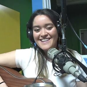 Natalia Valencia (a woman with long dark hair and a big smile, wearing headphones sitting at a microphone and playing guitar)