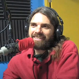 Neal Moogk-Soulis (a bearded man with long hair wearing a red shirt sits at a microphone)