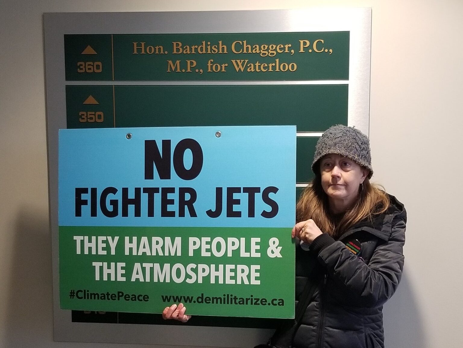 (Tamara Lorincz in front of a sign indicating "Hon. Bardish Chagger, P.C. | M.P. for Waterloo", holding a sign that reads "No Fighter Jets | They Harm People & The Atmosphere | #ClimatePeace www.demilitarize.ca)
