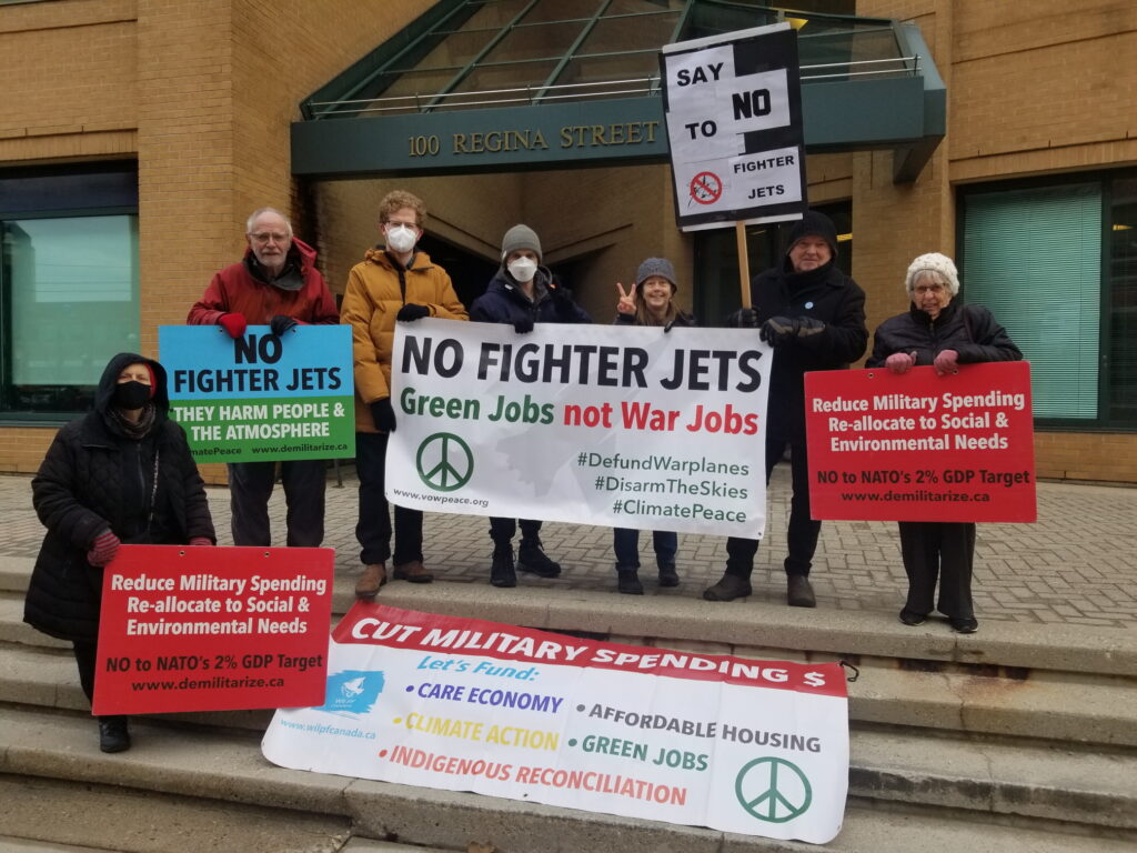 Protesters standing on the steps in front of the building at 100 Regina Street South holding signs with slogans such as #NoFighterJets, with one banner laid out on the steps