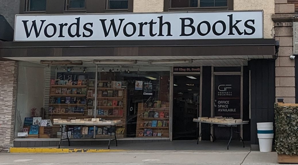 The storefront of Wordsworth Books in Uptown Waterloo with the front door open, two tables of books outside and three book display cases in the large front window.