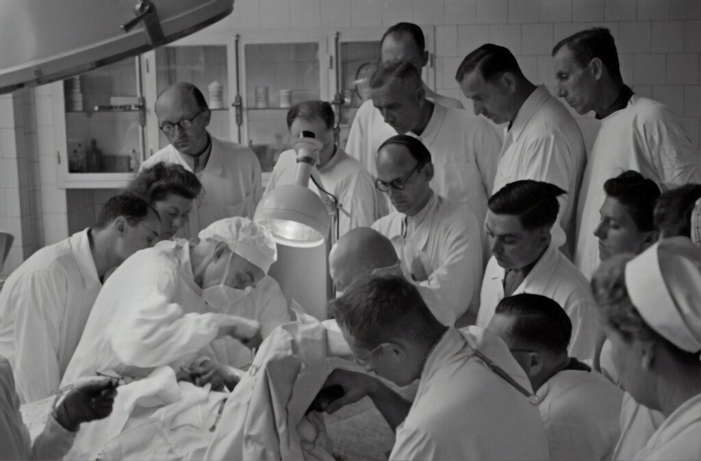 In a black and white photograph, a group of people, including doctors and nurses, stand in an operating room watching a doctor perform an operation. The clothes and hairstyles of the people in the room suggest the photo was taken in the mid-twentieth century.
