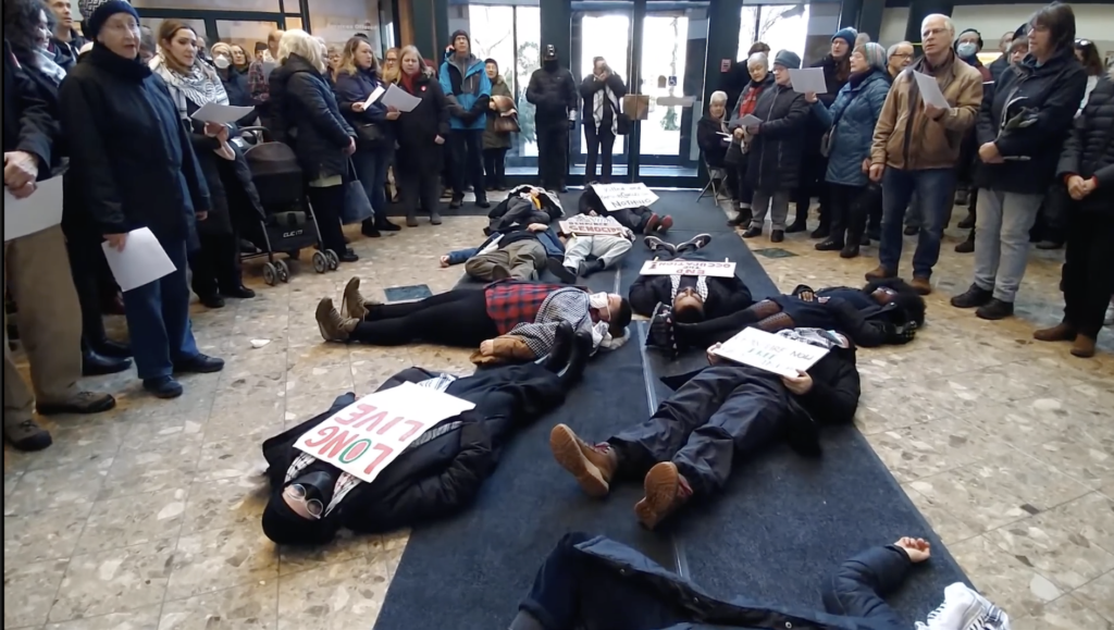 A group of people lie on the ground in the foyer at Waterloo City Hall pretending to be dead while another group stand around them singing hymns to draw attention to the need for a ceasefire between Israel and Palestine.