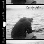 Reykjavictim (person sitting with back 3/4 to the camera, apparently with head in hands, in front of water; image framed by edges of film)