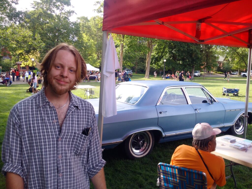 Riley Powis wearing a checkered shirt stands behind the CKMS-FM booth (only a corner of the red canopy is visible). Behind him is a person wearing a white baseball cap and yellow T-shirt, in the background is Riley's antique (1970s) blue car.