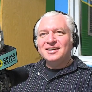 A man with white hair wearing headphones at a microphone with the CKMS-FM logo on it.