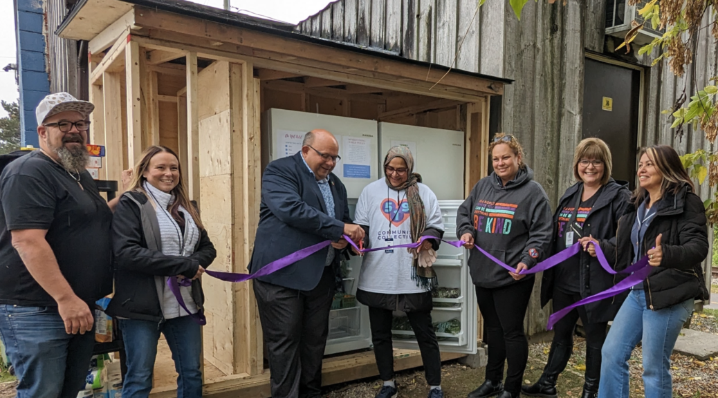 Seven people stand in front of the unfinished community shed building, cutting a ribbon to declare it open. Tyzun James, owner of Cafe Pyrus, and Kitchener mayor Berry Vrbanovic stand beside five board members of the 519 Community Collective as the cut the ribbon opening the new and improved Community Fridge at the Cafe Pyrus Outpost in Kitchener
