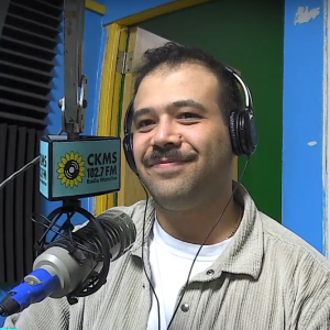 (A man with a moustache wearing a beige shirt and headphones smiles while sitting at a microphone)