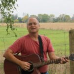 (Steve Todd playing acoustic guitar in a field)
