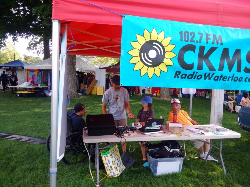 The CKMS-FM booth showing the table with a laptop and a speaker, four people standing or sitting behind the table. A CKMS-FM banner with the sunflower logo and the partial text "102.7FM CKMS RadioWaterloo.ca" is hanging from the red canopy.