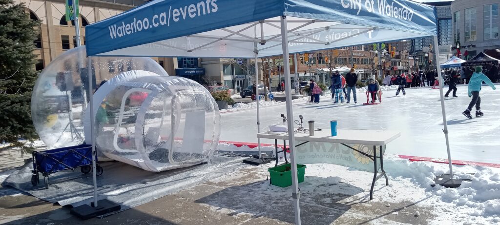 A blue canopy tent with "Waterloo.ca/events" and "City of Waterloo" on the edges; a table underneath holding two microphones and coffee mugs; a clear vinyl bubble dome for the DJ beside it, and a skating rink with lots of people skating in the background.