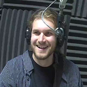 A smiling man wearing a blue shirt over a black T-Shirt and headphones sits at a microphone in front of  acoustic foam.
