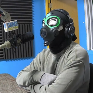 VEFO (a person with crossed arms sitting at a microphone wearing a gray sweater and grey gloves, and a gas mask over a Guy Fawkes mask; the gas mask has the eyes outlined in bright green paint, and there is a green heart painted on the forehead)