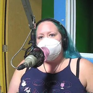 (a woman with blue hair wearing a purple dress and a facemask sits at a microphone)