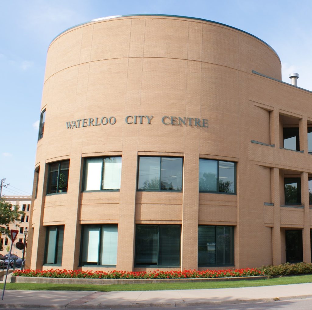 A picture of Waterloo City Centre, a leased building where city council meets and have their offices. The picture shows the front of the 3 story light brown brick building with windows and the words "Waterloo City Centre" on the building in a serif font. There are red flowers in a garden bed in the front of the building.