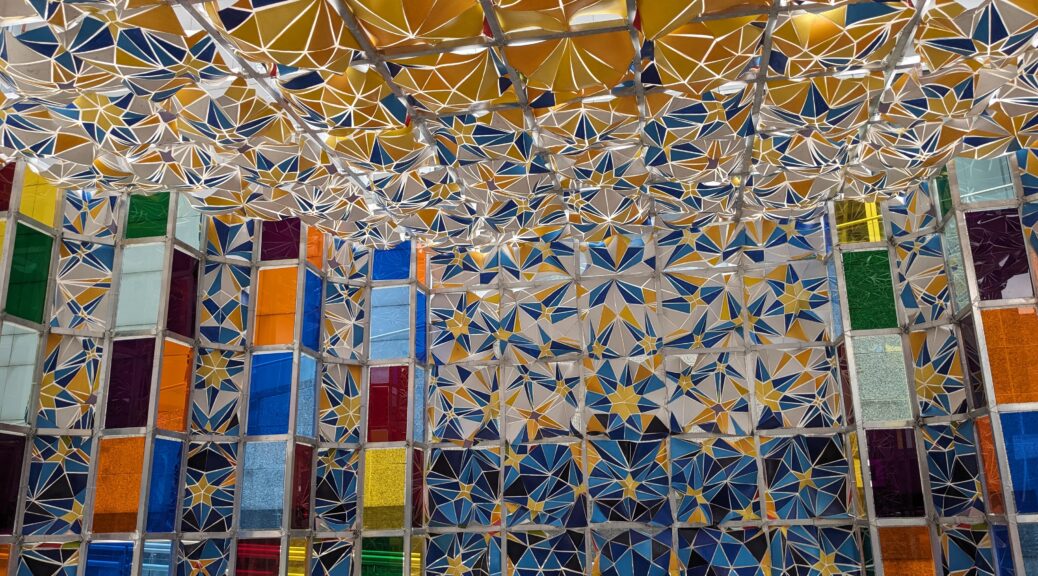 The picture shows the inside of a large artwork, with high vertical towers of panelled coloured glass and squares of fabric placed over a large steel structure and cut into a moqarnas pattern (a form of the ornamented vaulting used in Islamic architecture)