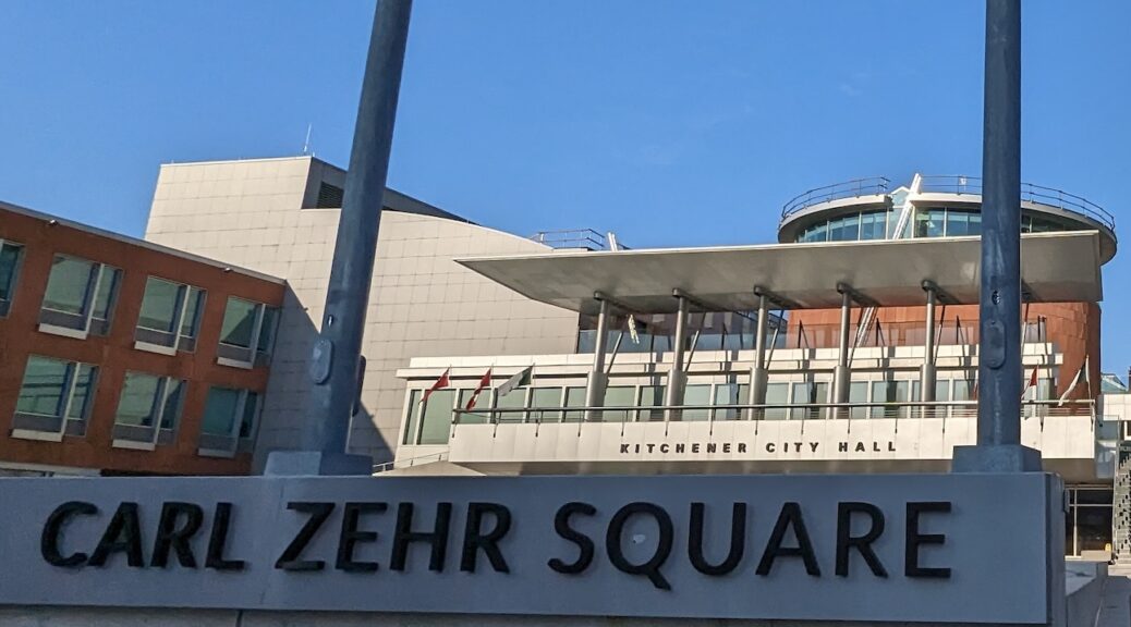 The second floor of a modern building taken from the sidewalk and looking up. A sign at the bottom of the frame says 'Carl Zehr Square'.
