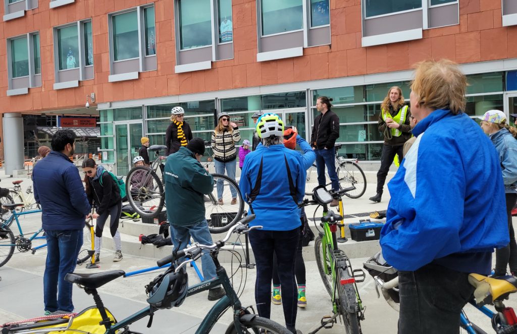 A picture from EarthDay In Kitchener-Waterloo. A group of people stand with their bikes, mingling, fixing, and listening to speeches from folks on a short stage. Waterloo Councillor Jen Vasic appears to be speaking. She has a sweater on. 1/2 the crowd has their bike helmets on.