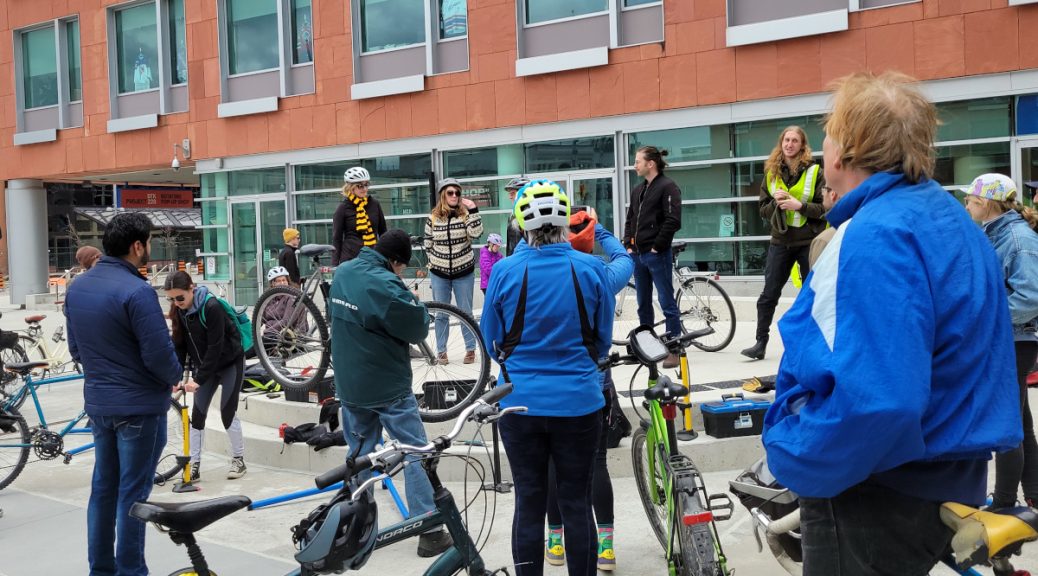 A picture from EarthDay In Kitchener-Waterloo. A group of people stand with their bikes, mingling, fixing, and listening to speeches from folks on a short stage. Waterloo Councillor Jen Vasic appears to be speaking. She has a sweater on. 1/2 the crowd has their bike helmets on.