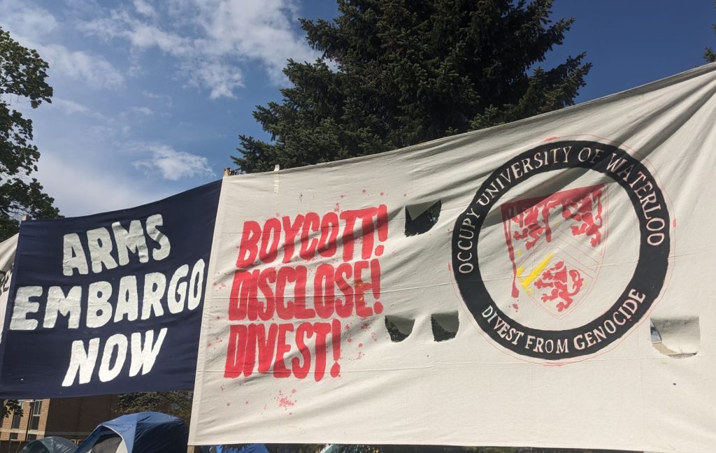 A close up picture of two banners with spruce trees and blue skies with a few clouds in the upper background. The dark blue banner has white text "Arms Embargo Now" and a white banner has the red text "Boycott! Disclose! Divest!" beside a reproduction of UWaterloo's logo, with the coat of arms in dripping red paint, and the circle around reading "Occupy University of Waterloo - Divest from Genocide" Tents are visible in the background beneath the banners.