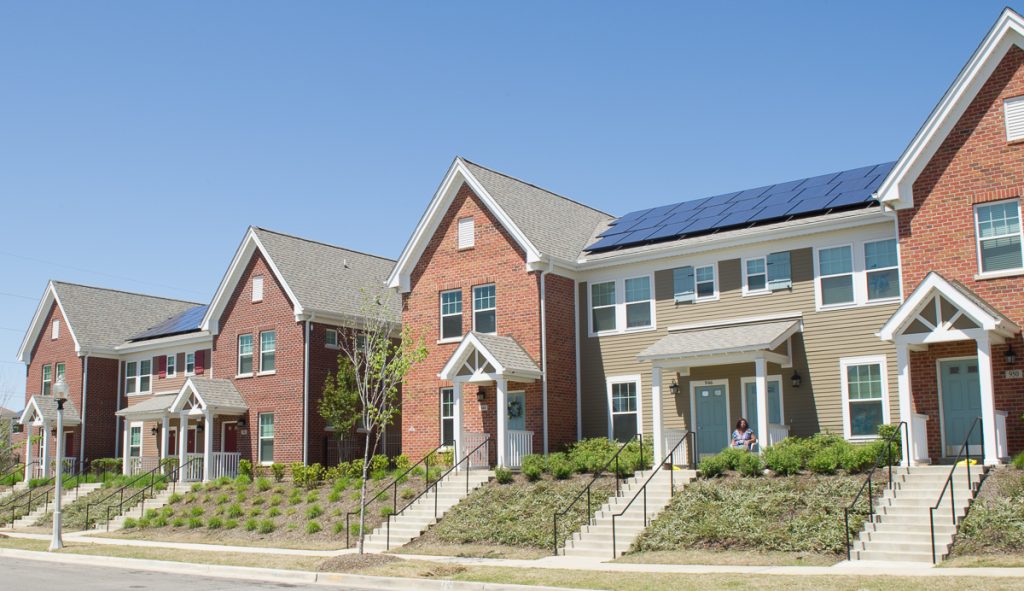 A picture of a row of townhomes in an affordable housing project in Memphis Tennessee with stairs leading to the doors, some simple pleasant landscaping with small shrubs in front of the homes. Homes are red brick and siding and have solar panels on the roof. Blue sky in the background.