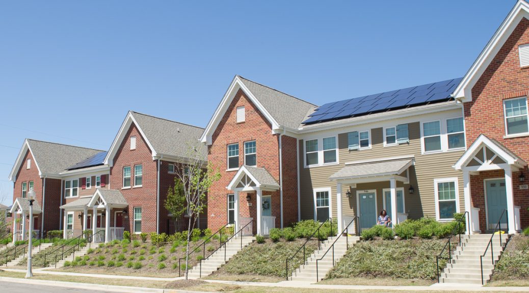 A picture of a row of townhomes in an affordable housing project in Memphis Tennessee with stairs leading to the doors, some simple pleasant landscaping with small shrubs in front of the homes. Homes are red brick and siding and have solar panels on the roof. Blue sky in the background.