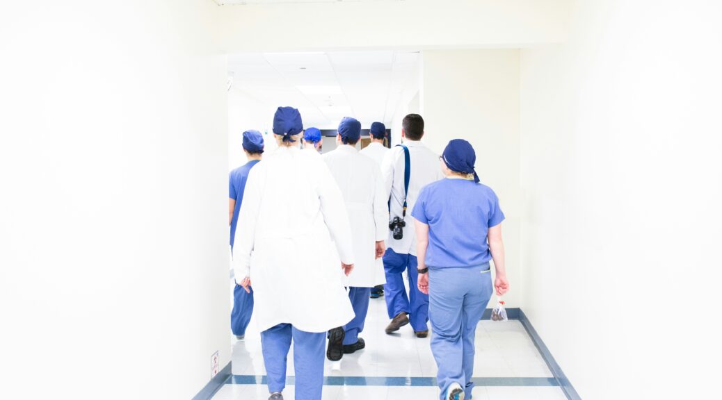 Seven medical professionals dressed in blue scrubs and white lab coats walk down a blinding white hallway away from the camera