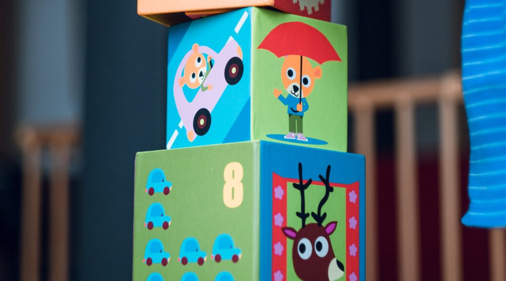 A child's hand steadys the top block of four blocks stacked on top of one another. The blocks have cartoon cariactures of a donkey, cats driving a car and holding an umbrella and a moose