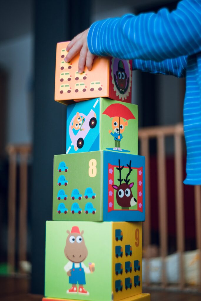 A child's hand steadys the top block of four blocks stacked on top of one another. The blocks have cartoon cariactures of a donkey, cats driving a car and holding an umbrella and a moose
