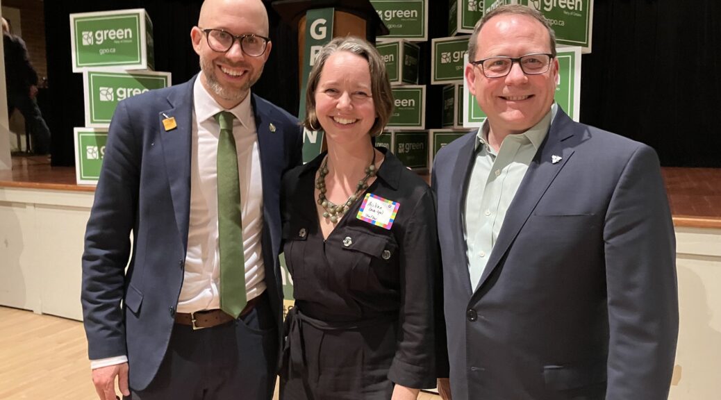 A colour photo of MP Mike Morrice, candidate Aislinn Clancy, and MPP Mike Schreiner standing in a tight line, smiling. They are all wearing business attire and standing infrom of "green Party" placards that are placed on a stage in the background.