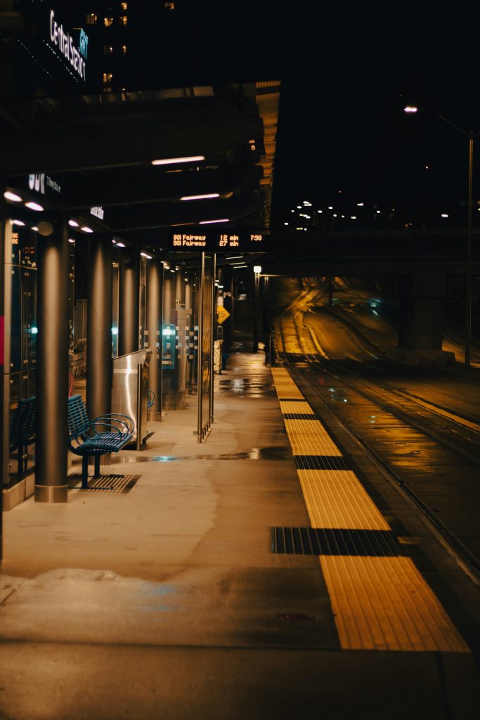 Warm light illuminating a platform at a train stop on a rainy dark night. The digital display notes how long until the next 2 trains arrive.