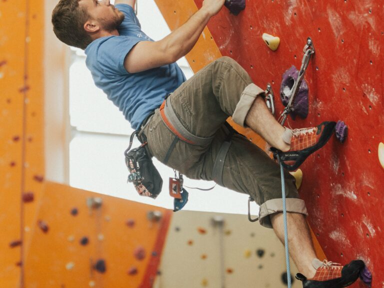 A man in a climbing harness climbs up an orange and red wall in a climbing gym