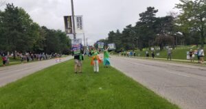 People and signs on both sides of a road, with pepole and signs in the grassy median as well. Signs reading "Stop The Sprawl" Among Others.