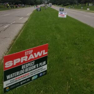 Lawn signs placed on either side of the road and in the median ahead of the Stop the Sprawl Rally. The first sign, red and dark green with white text which reads: "Stop Sprawl. Respect Waterloo Region's Plan. Stop Forced Urban Boundary Expansion on Farmland and Natural Spaces.