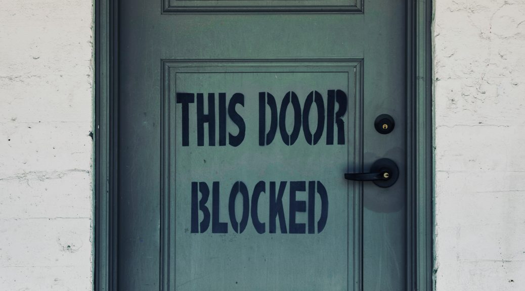 A light green door is inset into a brick wall; in the bottom panel of two on the door, it reads "This Door Blocked" in capital black letters