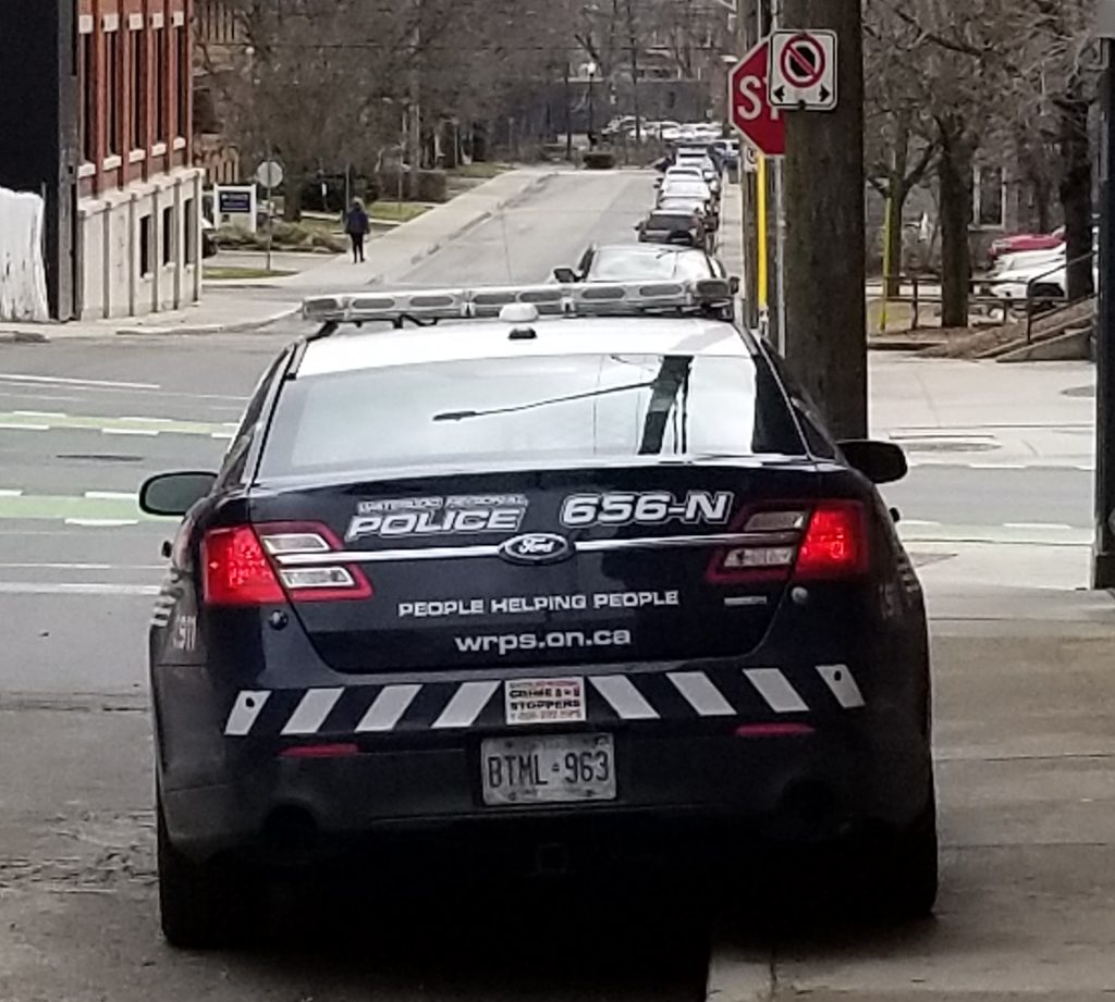 A photo of a spring day in Uptown Waterloo where a behind the car angle of a Waterloo Region police service cruiser parked on a the road AND the sidewalk out side of a doughnut and coffee shop, directly infront of a "No Stopping" sign. The motto "People Helping People" is visible on the trunk, along with the car's designation 656-N.